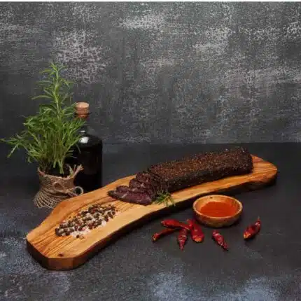 a piece of biltong slab with a few slices on a wooden board next to spices and herbs