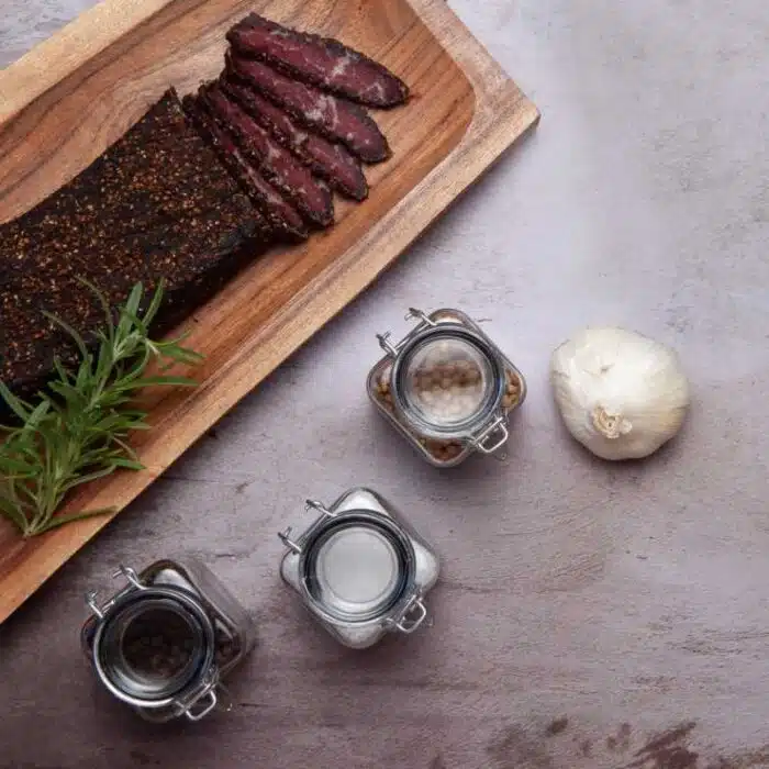 A sliced biltong slab on a wooden tray next to jars of spices