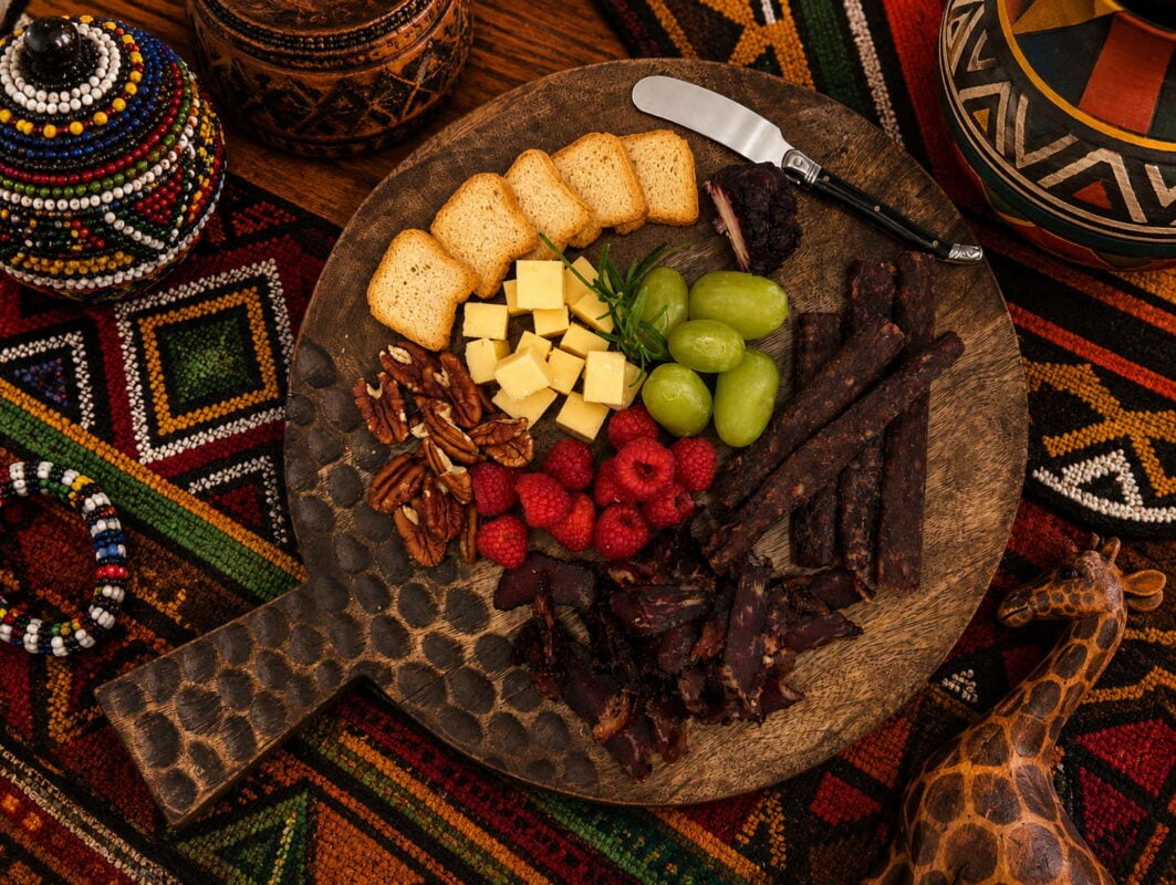 African charcuterie board with cheese, biltong, grapes, raspberries, and nuts on patterned fabric.