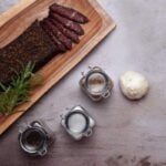 Rustic wooden board with seasoned beef jerky, rosemary, garlic, salt, and pepper jars.