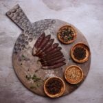 Rustic wooden charcuterie board with cured meat slices, assorted spices, herbs, and coarse salt.