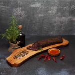 Rustic wooden board with sliced cured meat, spices, chili peppers, and fresh herbs.