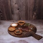 Rustic wooden board with sliced grilled steak, sea salt, peppercorns, and assorted spices.