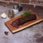 Rustic wooden board with sliced spiced cured meat, herbs, garlic, and seasoning jars.